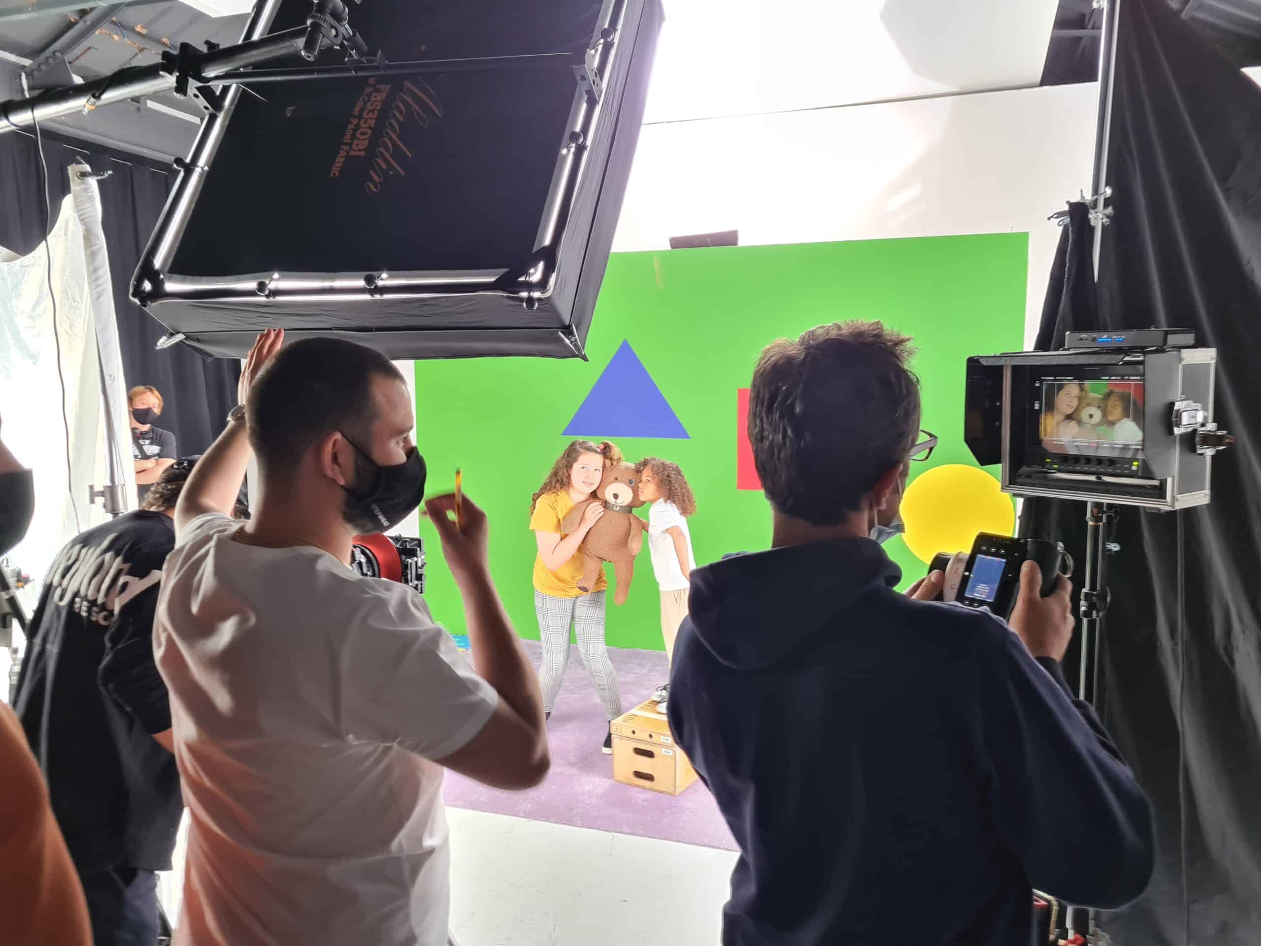 A behind-the-scenes image of the Twisted Toys campaign. Three men are in the foreground observing the camera or set. There is lighting equipment and cameras positioned towards a set that features two girls holding a teddy bear with a camera in its left eye. They are stood on a purple carpet, with a bright green backdrop that features a blue triangle and red square.