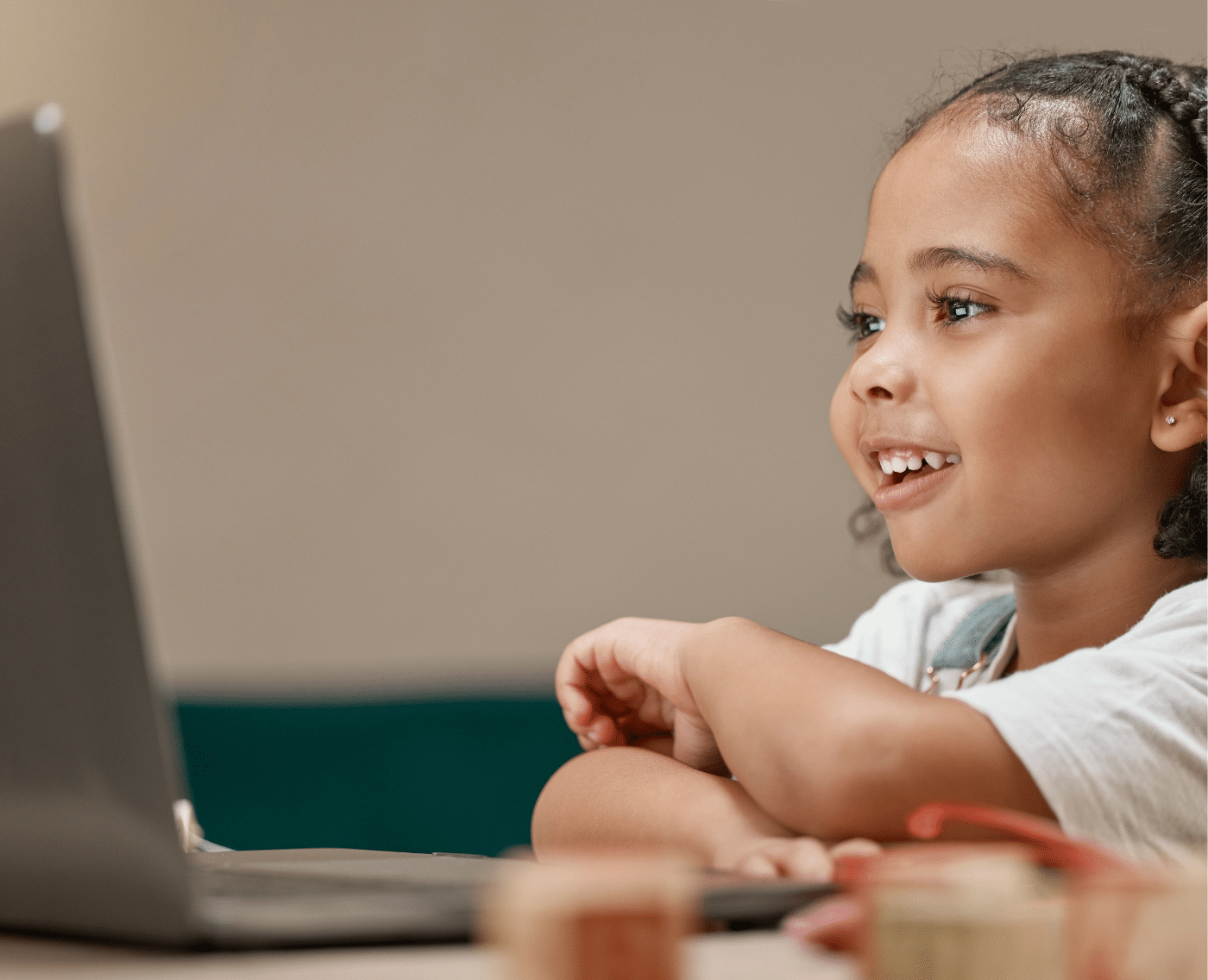 Young black girl on a laptop. Smiling and looking curiously at a laptop. Both the girl and the laptop are side on. The laptop is to the left of the image, and the girl is to the right. 