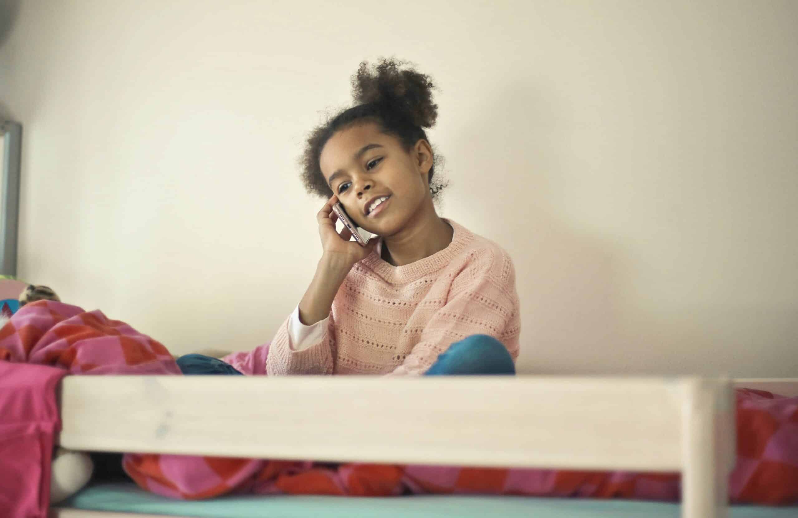 A black girl, sat on her bed on top of her duvet. She is holding a smartphone in right hand and holding it to her ear. Her head is tilted to the left and she is smiling.