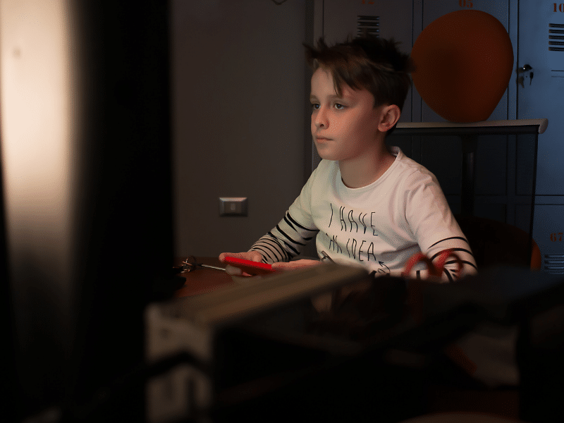 A young boy with short hair intently looks at a computer screen in a dimly lit room with lockers in the background.