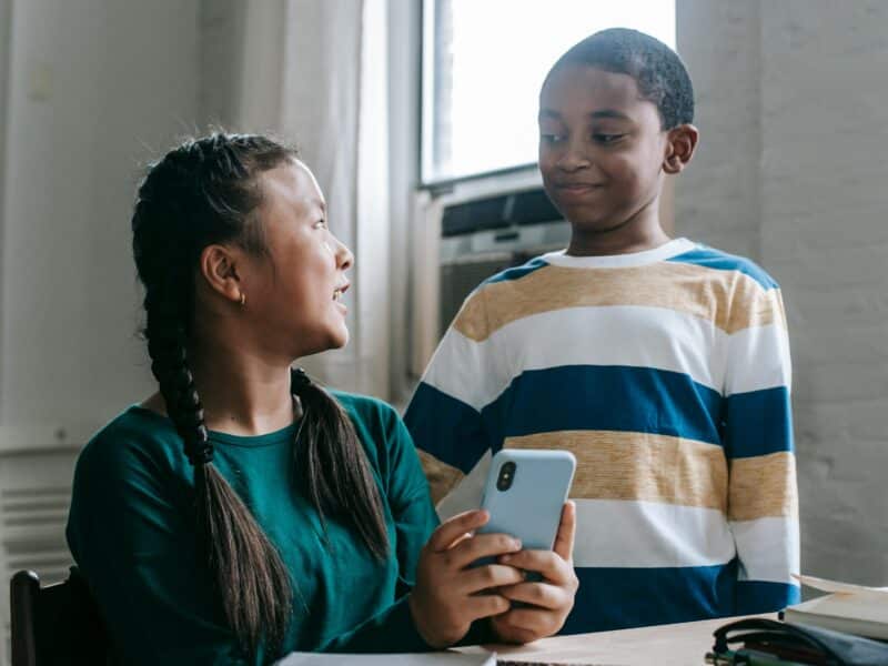 A young girl with black hair pleated in braids and wearing a teal long-sleeve shirt is showing something on her phone to her friend, a boy seemingly younger than her wearing a striped long-sleeve shirt. The boy is standing while she is sitting down.