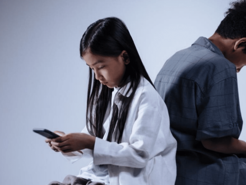 Two children, a girl and a boy, are sitting back-to-back while both looking down at their smartphones. The girl has long black hair and is wearing a white sweater. The boy has short curly hair and is wearing a gray shirt. The background is plain and light-coloured.