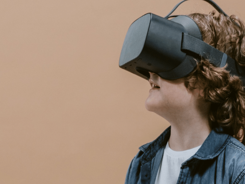 A young child with curly hair is wearing a virtual reality headset and smiling. They are dressed in a denim jacket and a white shirt, standing against a plain beige background.