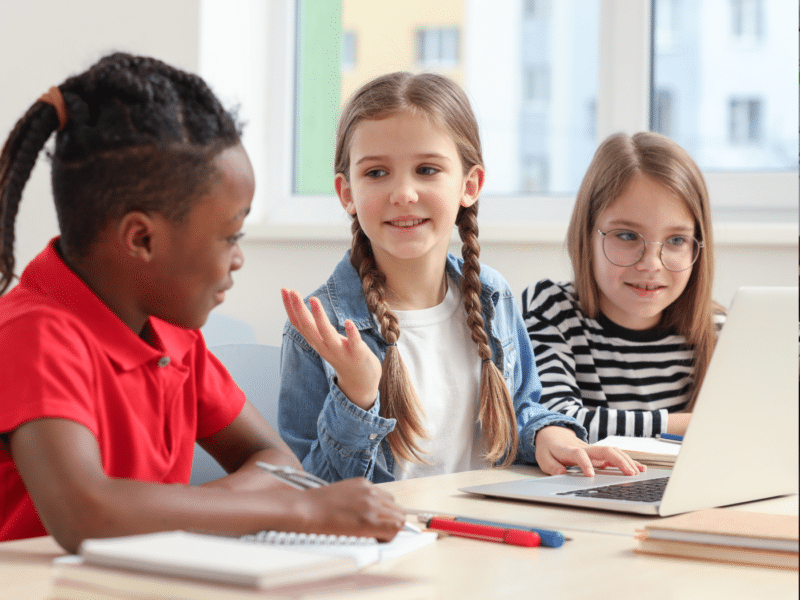 Three children are sitting at a table, engaged in a group activity. The child on the left is wearing a red shirt and has a notebook in front of them. The child in the middle, with braids, is gesturing and is sitting in front of a laptop while the child on the right, in glasses, is looking intently at the laptop screen.