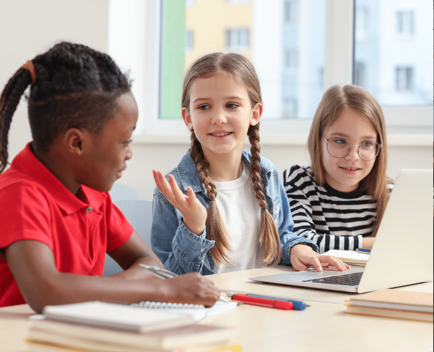 Third young children are sat at a desk on a laptop. The black boy on the left is wearing a red polo shirt. The white girl in the middle is looking and speaking to the black boy, gesturing with her right hand, with her left hand placed on the laptop's trackpad. The white girl on the right has round glasses and is looking at the laptop screen. The image is bright and suggests a friendly atmosphere amongst the children.