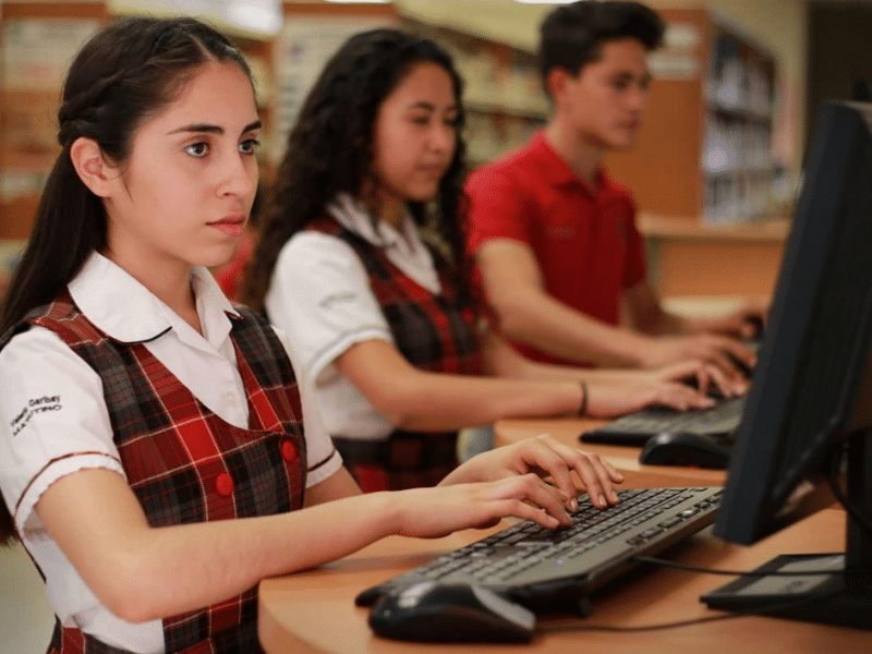 Three students in school uniforms work at computers in a library. Shelves filled with books are visible behind them.