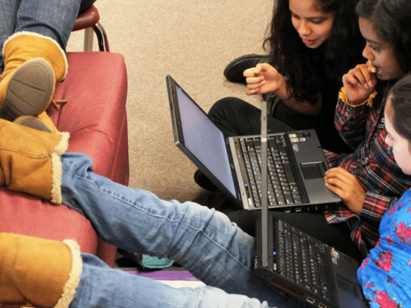 Three children sit close together, each focused on their own laptops. The child on the left is reclined with their legs extended on a chair, wearing brown winter boots. The children appear to be working collaboratively or engaged in their individual projects.