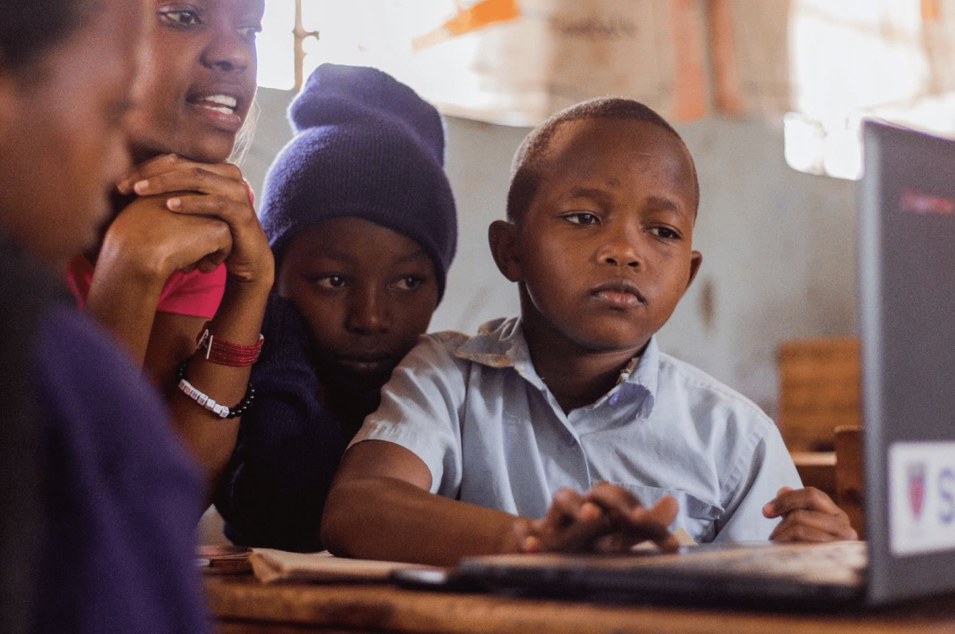 Four black children are looking at a laptop in a classroom, and one of them is touching the trackpad.