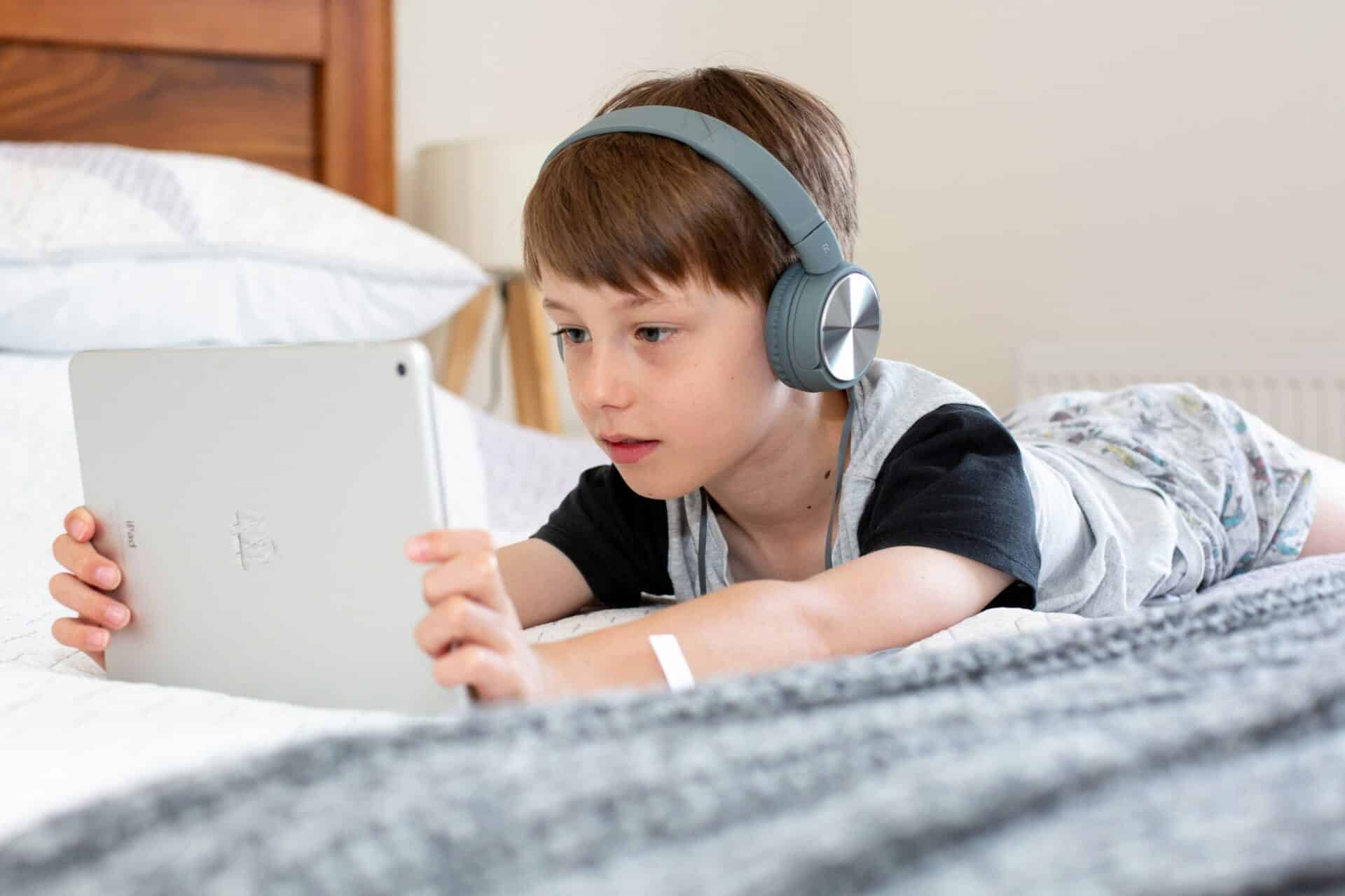 A young boy wearing headphones lying on a bed holding his tablet device.