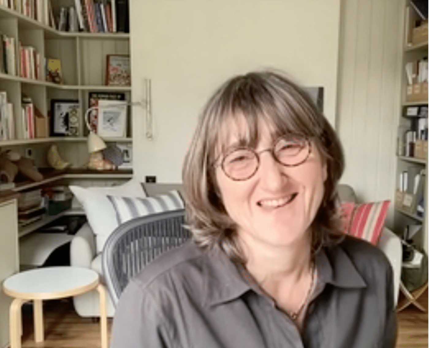 A photo of Baroness Beeban Kidron smiling warmly as she sits in a well-lit room. She is wearing round glasses and a dark blouse, with her hair framing her face. The background shows bookshelves filled with books and decorative items, a striped cushion on a sofa, and a lamp. This image captures the moment when she accepted the EPIC 2023 Champion of Freedom award, recognising her pioneering work on children's online privacy and the Age-Appropriate Design Code.