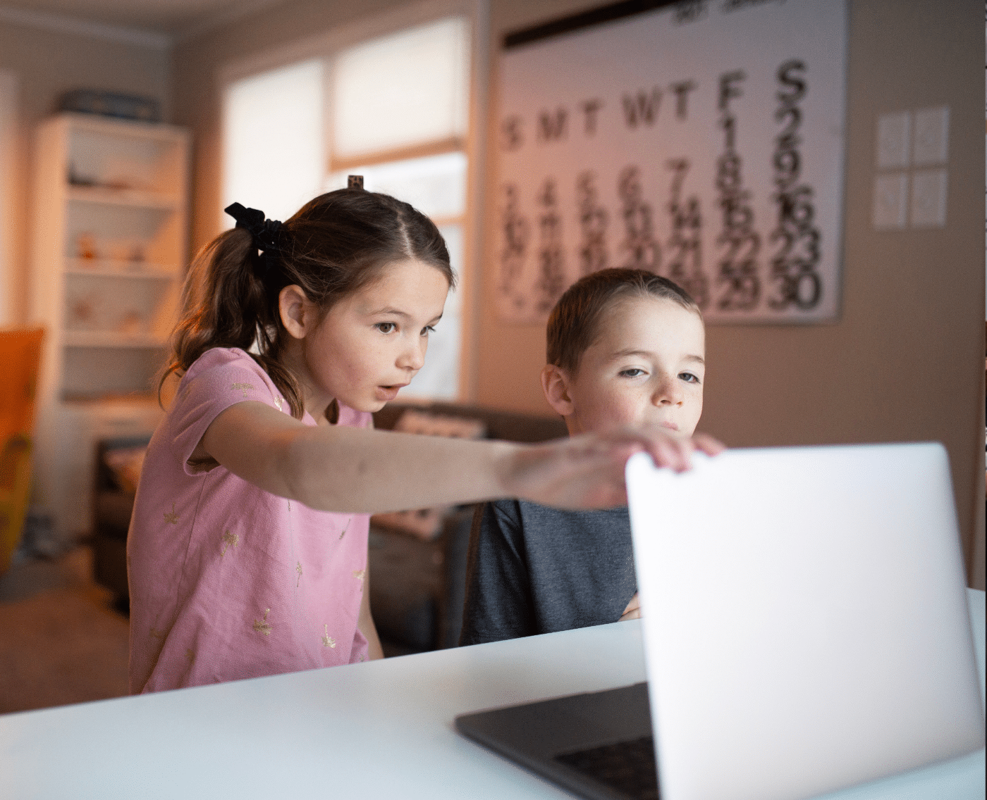 Two children are sitting at a table, focused on a laptop. The girl points at the screen while the boy looks on attentively. The background shows a cosy room with a yellow chair, a bookshelf, and a large calendar hanging on the wall.