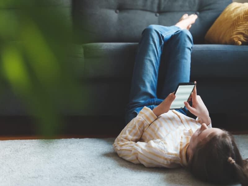 Teen girl with smartphone relaxing at home. She is lying on the floor, with legs resting on the sofa. There is an out-of-focus plant in the forground, to the left of the image.