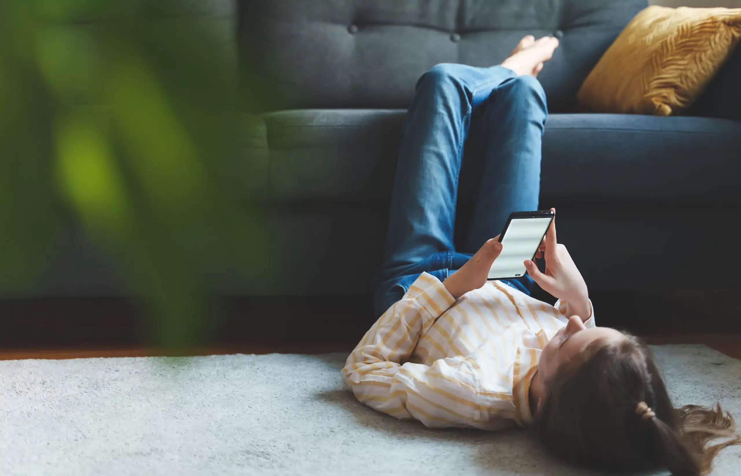 Teen girl with smartphone relaxing at home. She is lying on the floor, with legs resting on the sofa. There is an out-of-focus plant in the forground, to the left of the image. 
