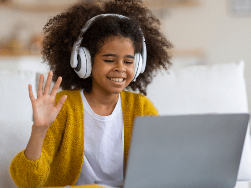 A young black girl with curly brown hair wears large headphones while sitting on a white couch. She is smiling and waving at a laptop screen in front of her, indicating she is likely attending an online class. She is dressed casually in a mustard-yellow cardigan over a white t-shirt. She is sitting in a well-lit living room with neutral tones in the background, and a notebook is partially visible on the table in front of her.