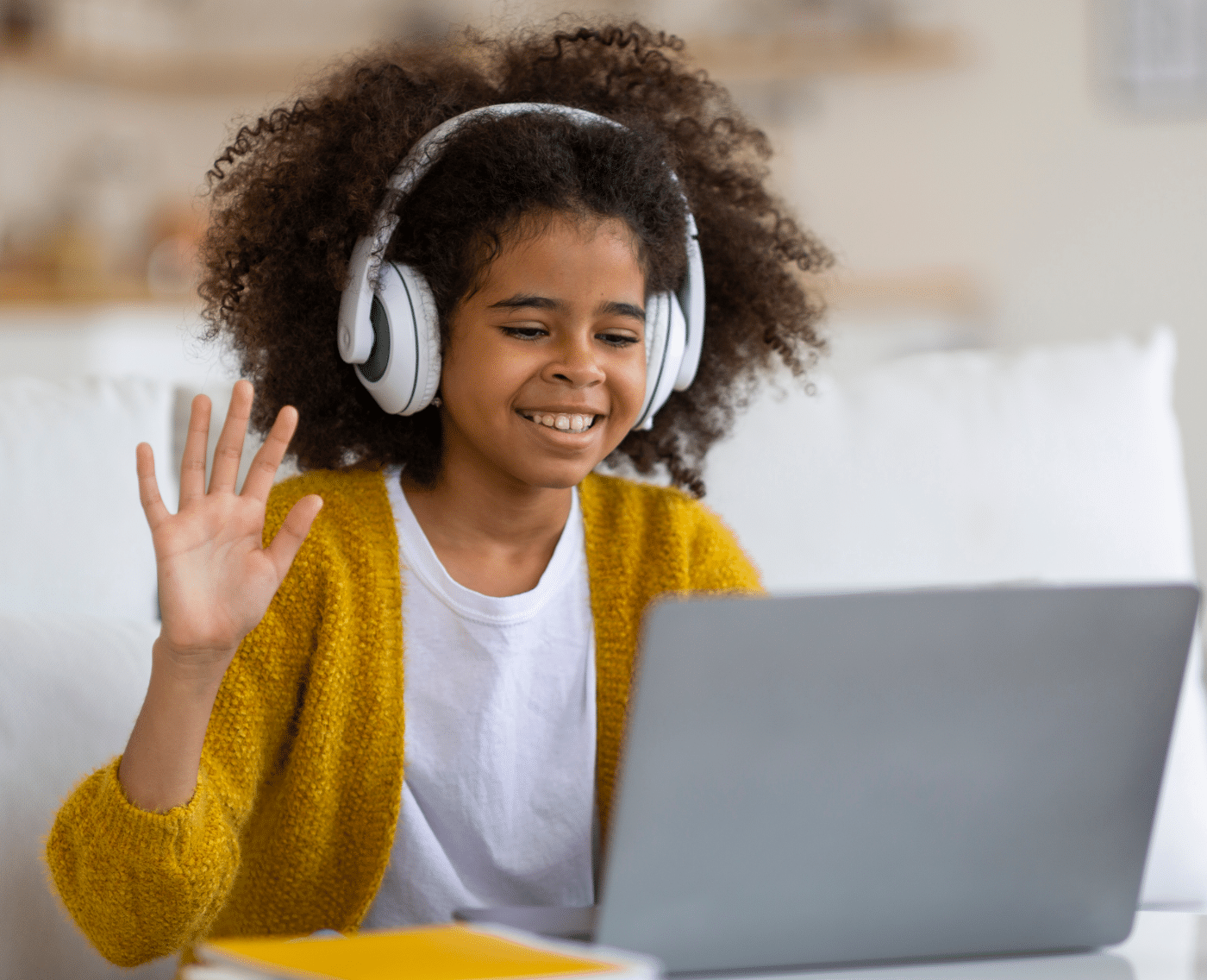 A young black girl with curly brown hair wears large headphones while sitting on a white couch. She is smiling and waving at a laptop screen in front of her, indicating she is likely attending an online class. She is dressed casually in a mustard-yellow cardigan over a white t-shirt. She is sitting in a well-lit living room with neutral tones in the background, and a notebook is partially visible on the table in front of her.