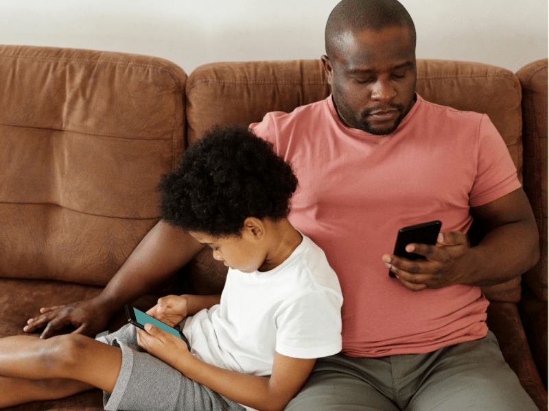 A father and his young son sitting together on a brown couch, both focused on their smartphones. The father, wearing a pink T-shirt and grey shorts, is holding his phone with one hand, while the son, dressed in a white T-shirt and grey shorts, leans against him, deeply engrossed in his own device.