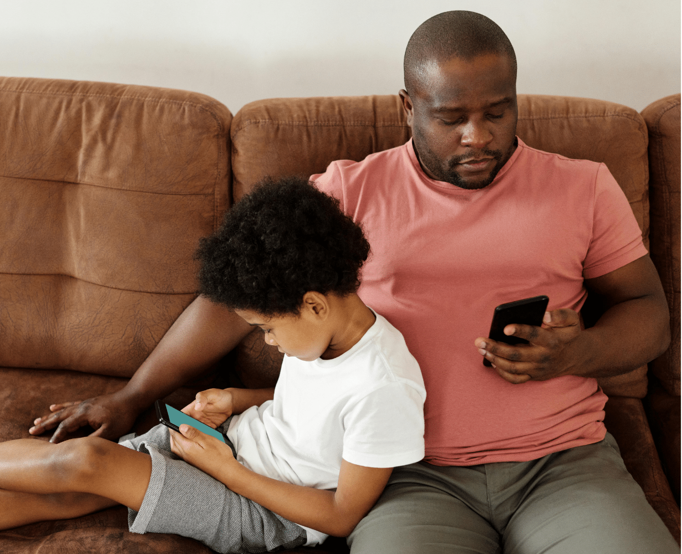 A father and his young son sitting together on a brown couch, both focused on their smartphones. The father, wearing a pink T-shirt and grey shorts, is holding his phone with one hand, while the son, dressed in a white T-shirt and grey shorts, leans against him, deeply engrossed in his own device.