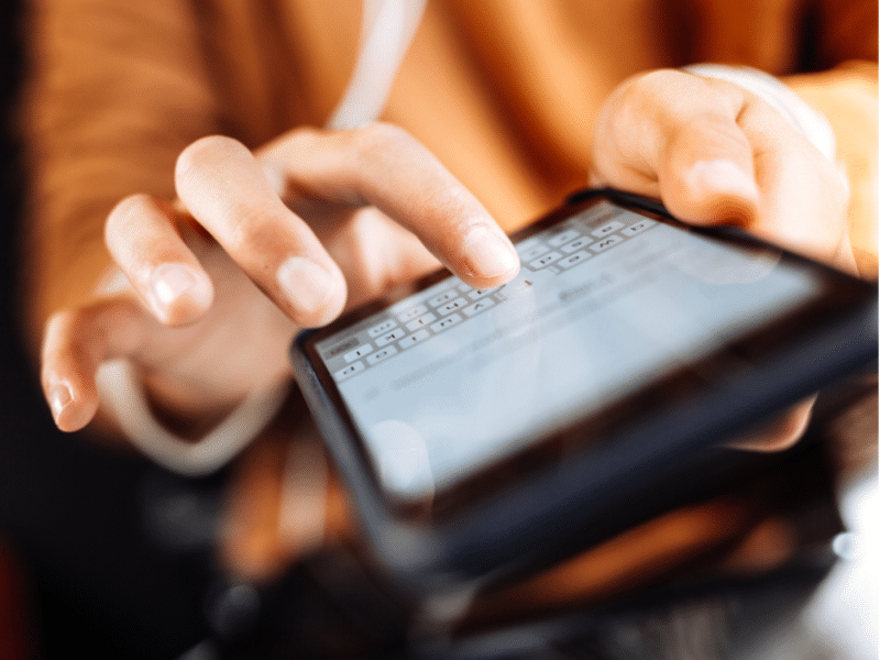 A close-up image of a person's hands typing on a smartphone. The fingers are in motion, interacting with the on-screen keyboard, which is slightly blurred. The person appears to be focused on the device, with the screen displaying text input. The background is blurred, drawing attention to the hands and the phone.