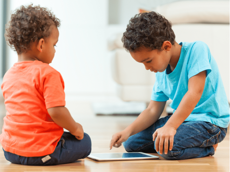 Two young children sitting on the floor of a bright, modern room, focused on a tablet device between them. The child on the left, wearing an orange shirt and dark pants, is sitting cross-legged and watching the screen intently. The child on the right, wearing a blue shirt and jeans, is leaning forward slightly, using one hand to interact with the tablet.