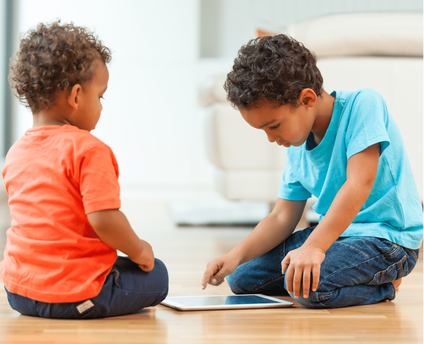  Two young children sitting on the floor of a bright, modern room, focused on a tablet device
between them. The child on the left, wearing an orange shirt and dark pants, is sitting cross-legged and
watching the screen intently. The child on the right, wearing a blue shirt and jeans, is leaning forward slightly,
using one hand to interact with the tablet.