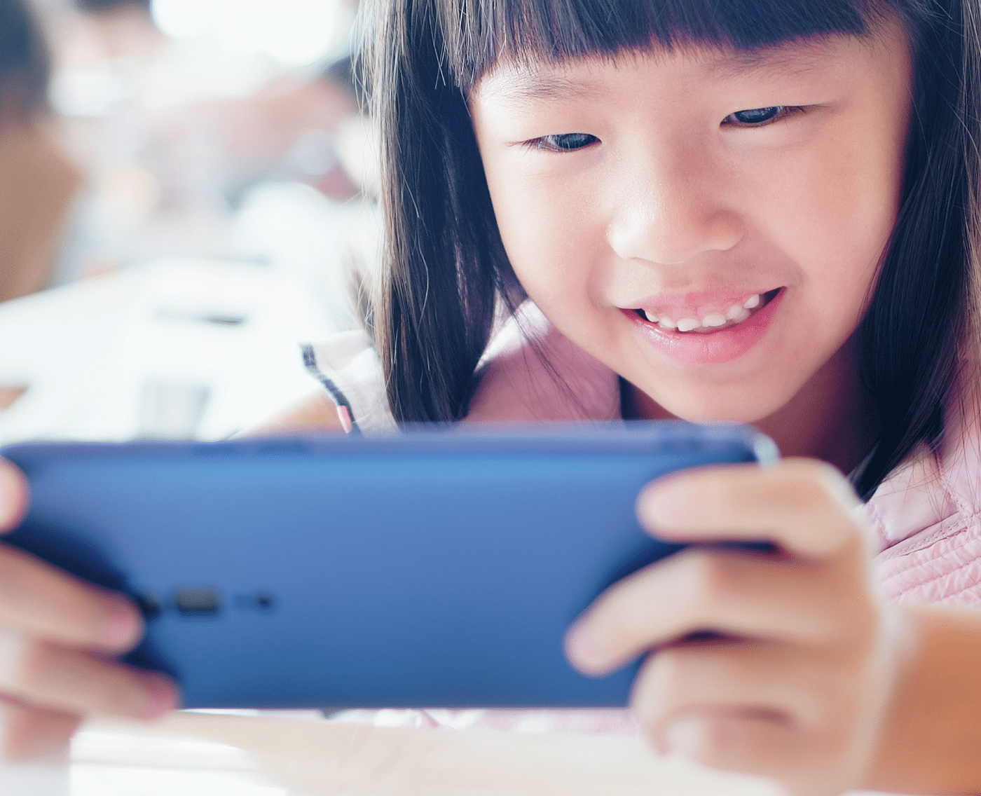 A young girl with straight black hair is smiling as she looks at a smartphone she is holding with both hands. The focus is on her face and the phone, suggesting she is enjoying a game or video. The background is blurred.