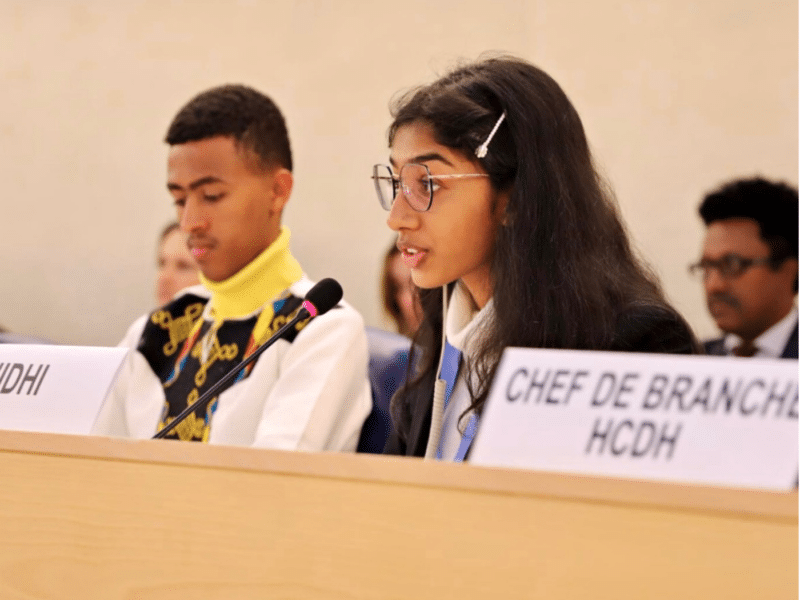 A young girl named Nidhi is speaking at a microphone during a session at the United Nations Human Rights Council. She is wearing glasses and a formal outfit, with her long dark hair neatly pinned back with a hair clip. Next to her is a young boy, also dressed formally, listening attentively.