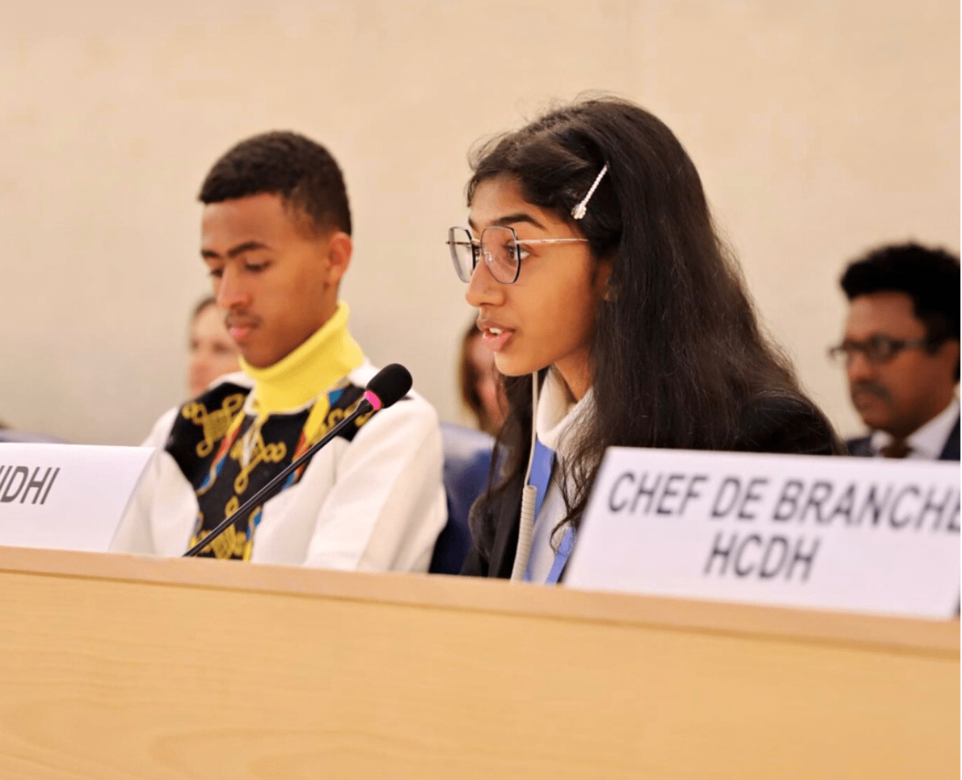 A young girl named Nidhi is speaking at a microphone during a session at the United Nations Human Rights Council. She is wearing glasses and a formal outfit, with her long dark hair neatly pinned back with a hair clip. Next to her is a young boy, also dressed formally, listening attentively.