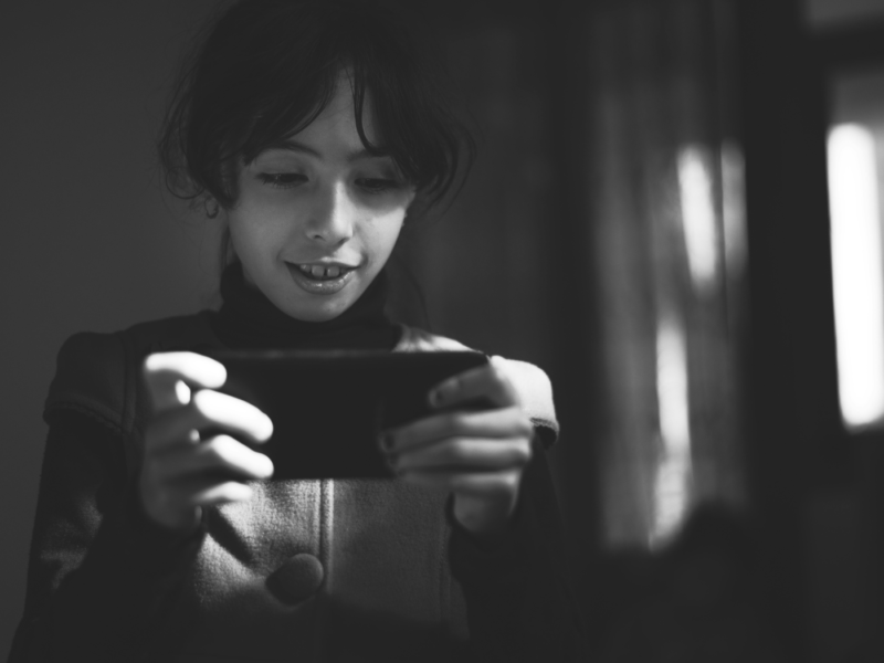 A black-and-white image of a young girl with dark hair, holding and looking at a smartphone with a focused expression. The lighting creates a soft contrast, highlighting her face and the device while the background remains dim and blurred. The girl appears to be engaged with the content on her phone, in a quiet and reflective moment.