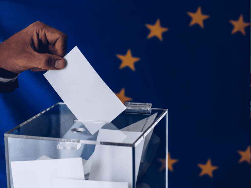 A person in a blue suit is dropping a white ballot into a clear ballot box. The person's hand is reaching into the frame from the left side of the image and their ballot is folded in half. The ballot box is made of glass and is sitting on a table in front of the flag of the European Union: a blue flag with twelve gold stars arranged in a circle.