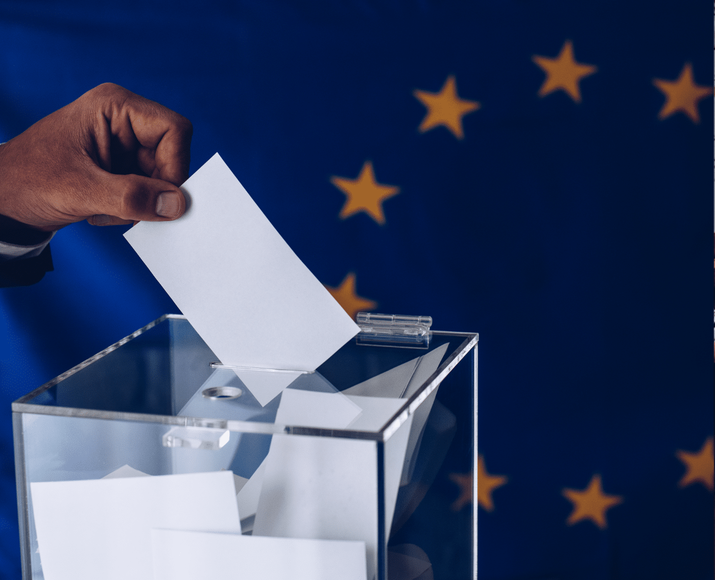 A person in a blue suit is dropping a white ballot into a clear ballot box. The person's hand is reaching into the frame from the left side of the image and their ballot is folded in half. The ballot box is made of glass and is sitting on a table in front of the flag of the European Union: a blue flag with twelve gold stars arranged in a circle.