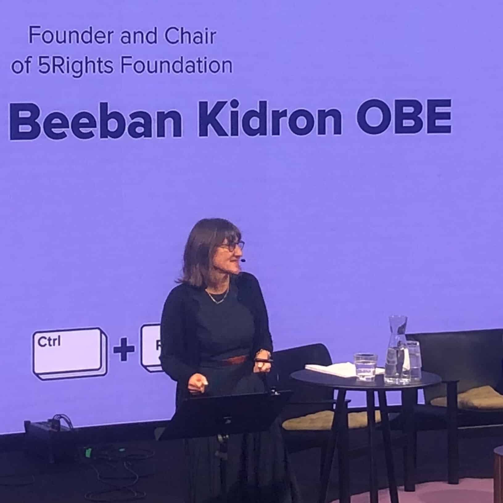 Baroness Beeban Kidron is speaking to an audience at the Ctrl + Rights Conference in Stockholm. She has dark straight hair and she is wearing black dress with a black cardigan. Behind her, the screen is lilac and showcases the logo of the conference – two keys from a keyboard, one saying Ctrl and one saying Rights. The screen also shows her full name.