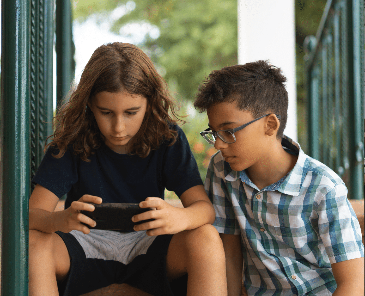 Two children sitting close together on a porch or outdoor steps, with one of them focused on a smartphone. The child on the left, who has long brown hair and is wearing a navy blue t-shirt, is intently looking at the phone he is holding in both hands. The child on the right, who has short dark hair and is wearing glasses and a green and white plaid shirt, is sitting slightly behind and to the side, also looking at the phone. The background shows some greenery, suggesting they are outdoors on a porch or patio. Both children appear to be concentrating on whatever is displayed on the phone, suggesting that they might be playing a game or watching something together.