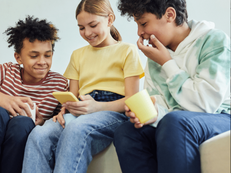 Three children sitting close together on a light-coloured couch. The child in the centre is holding a phone with a yellow case, while the other two children look on with interest and amusement. The child on the left is wearing a striped red and white t-shirt and has short curly hair. He is smiling softly as he looks at the phone. The child in the middle, who is holding the phone, is wearing a yellow t-shirt and has her hair tied back. She is smiling as she shows something on the screen to the others. The child on the right, wearing a white hoodie with green tie-dye patterns and dark pants, is laughing with his hand covering his mouth. He also holds a yellow cup in his other hand.