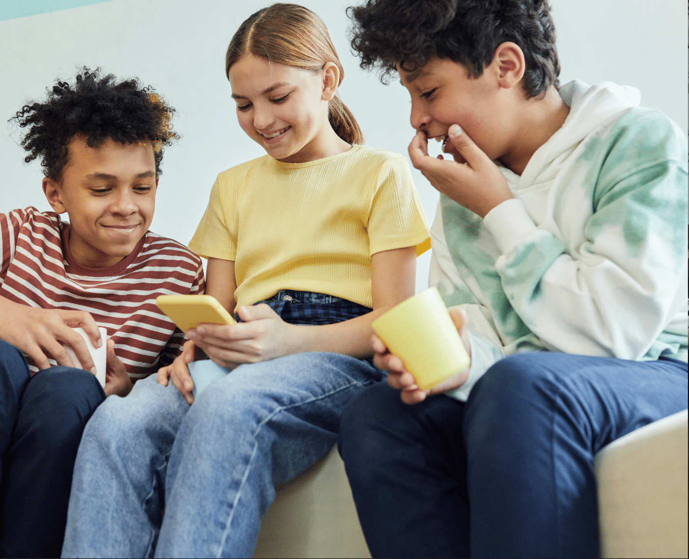 Three children sitting close together on a light-coloured couch. The child in the centre is holding a phone with a yellow case, while the other two children look on with interest and amusement. The child on the left is wearing a striped red and white t-shirt and has short curly hair. He is smiling softly as he looks at the phone. The child in the middle, who is holding the phone, is wearing a yellow t-shirt and has her hair tied back. She is smiling as she shows something on the screen to the others. The child on the right, wearing a white hoodie with green tie-dye patterns and dark pants, is laughing with his hand covering his mouth. He also holds a yellow cup in his other hand.