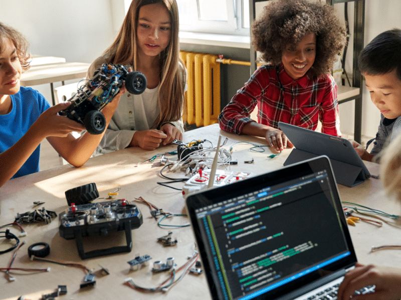 A diverse range of children are sat at a desk, engaging with robotic equipment and technology. They are happy and concentrating.