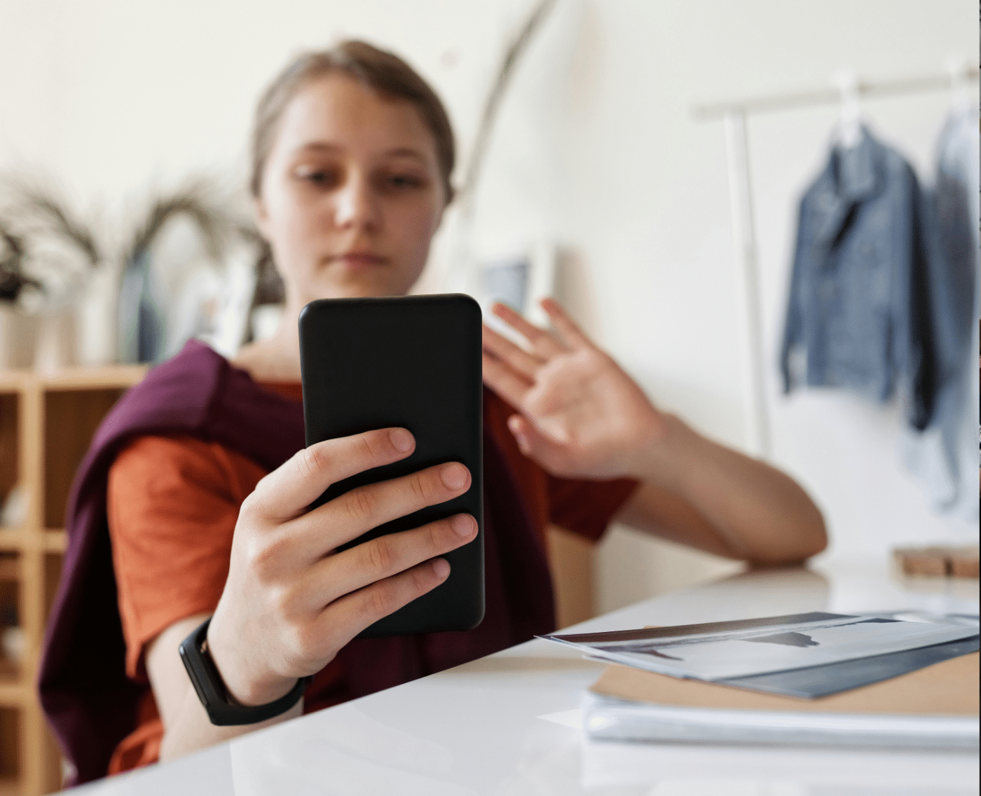 A young person is sitting indoors at a desk and holding a smartphone in their right hand. They appear to be engaged in a video call, as they are looking at the phone's screen and raising their left hand, possibly waving. The person is wearing an orange shirt and has a dark smartwatch on their left wrist. In the background, there is a shelf with potted plants and some hanging clothing, which is slightly out of focus, suggesting the photo was taken in a home or casual setting.