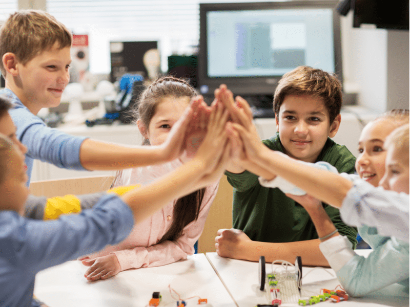 A group of young children, boys and girls, are gathered around a table in a classroom, enthusiastically giving each other a high-five. They are smiling and appear to be celebrating. The classroom environment is bright and modern, with a computer screen and other educational equipment visible in the background.