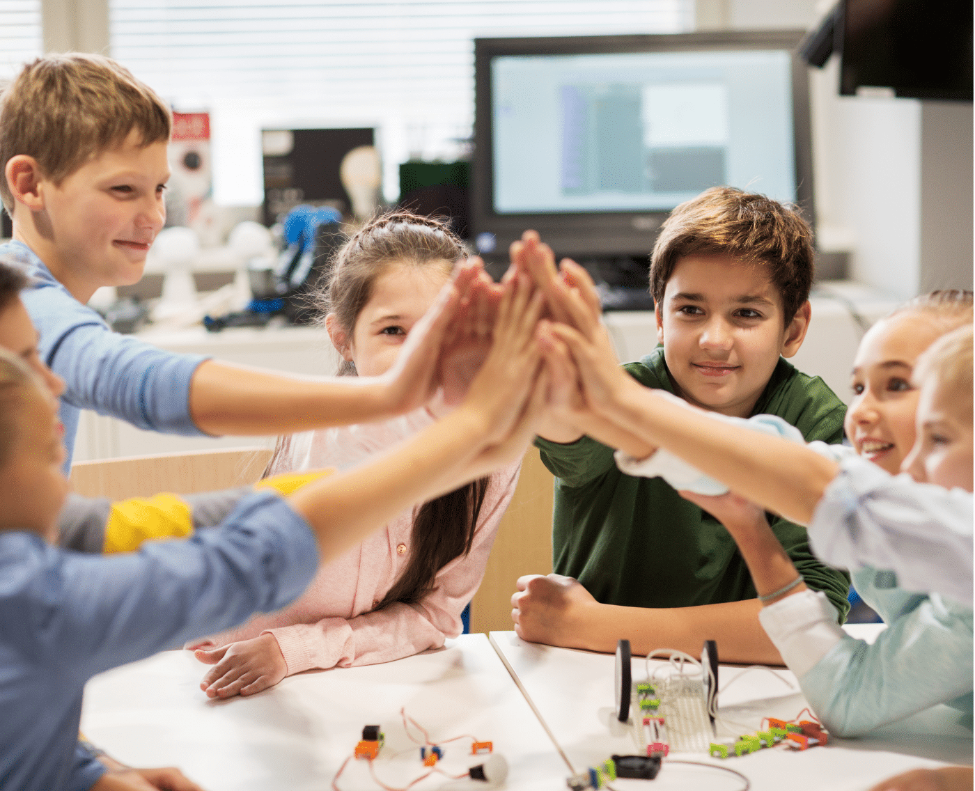 A group of young children, boys and girls, are gathered around a table in a classroom, enthusiastically giving each other a high-five. They are smiling and appear to be celebrating. The classroom environment is bright and modern, with a computer screen and other educational equipment visible in the background.