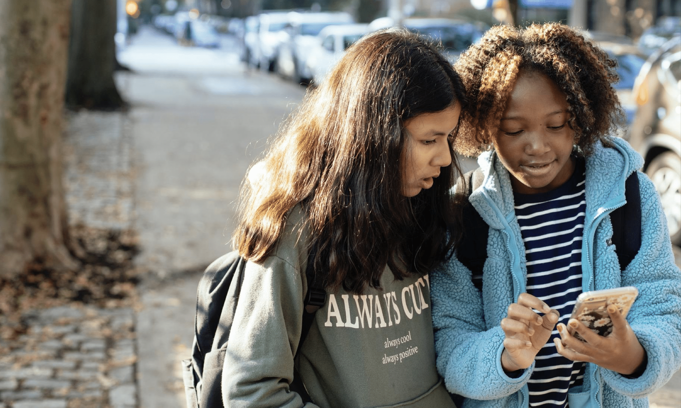 Two young girls are looking over a smartphone while smiling.