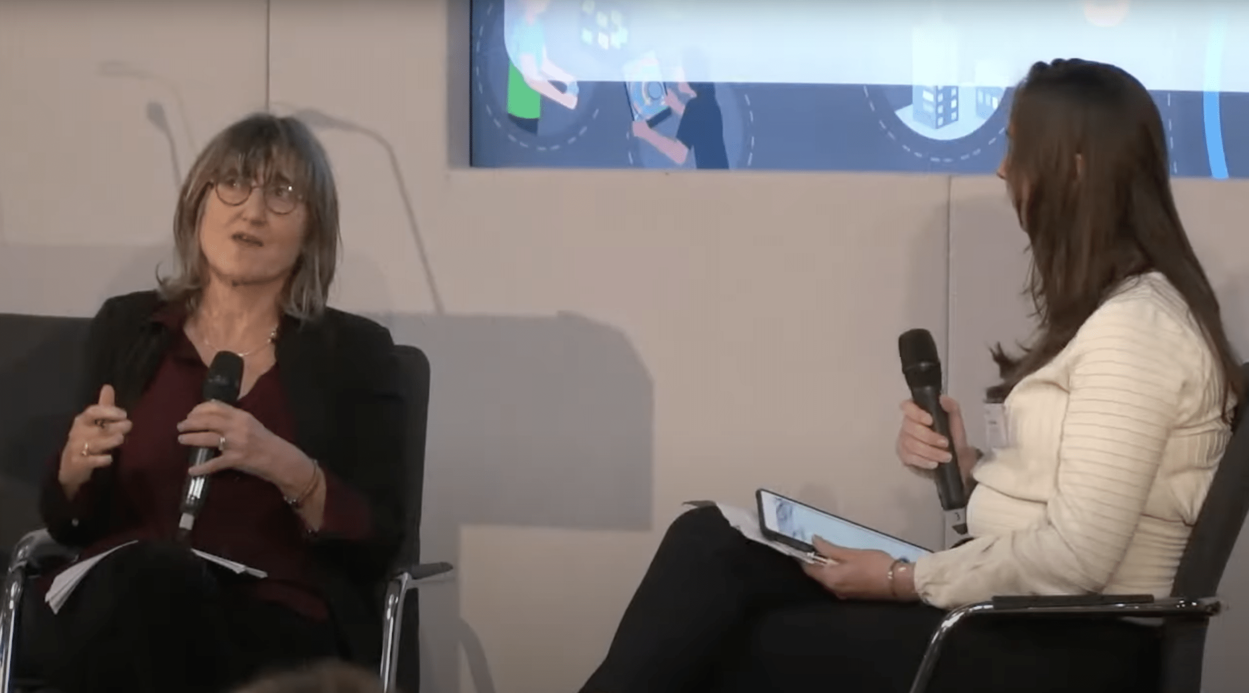 Baroness Beeban Kidron (on the left) participating in a Q&A session at an event organised by Queen Mary University in London, titled “AI at a Turning Point: How Can We Create Equitable AI Governance Futures?” She is seated, holding a microphone, and appears to be engaged in conversation, gesturing with her hand. Baroness Kidron is wearing glasses, a dark blazer, and a burgundy top. Across from her, another woman (on the right) is also seated, holding a microphone and a tablet, listening attentively to Baroness Kidron.