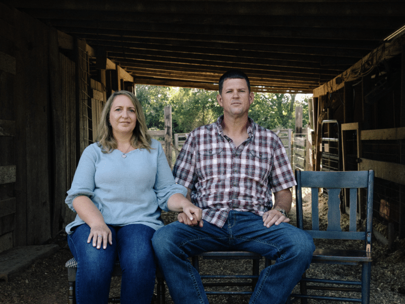 A woman on the left and a man in the middle. Both are sat on wooden chairs. They are positioned underneath a wooden stable. You can the trees and evening light towards the back of the stable. To the right of the man is an empty chair, used in this campaign to signify the child 'lost' to social media.