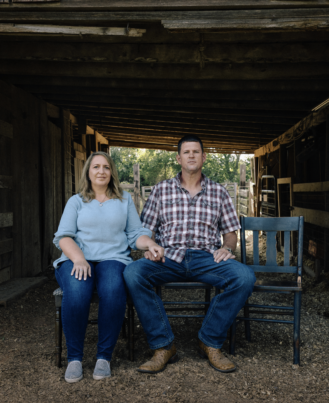 A woman on the left and a man in the middle. Both are sat on wooden chairs. They are positioned underneath a wooden stable. You can the trees and evening light towards the back of the stable. To the right of the man is an empty chair, used in this campaign to signify the child 'lost' to social media. 