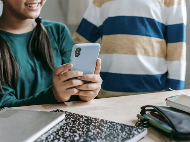 A girl is sitting at a desk, holding a smartphone. The desk is in the foreground. There are two notepads on the desk to the left of the image. A boy is standing to the girl's left. Both faces of the children are cropped out, but you can see the girl visibly smiling.