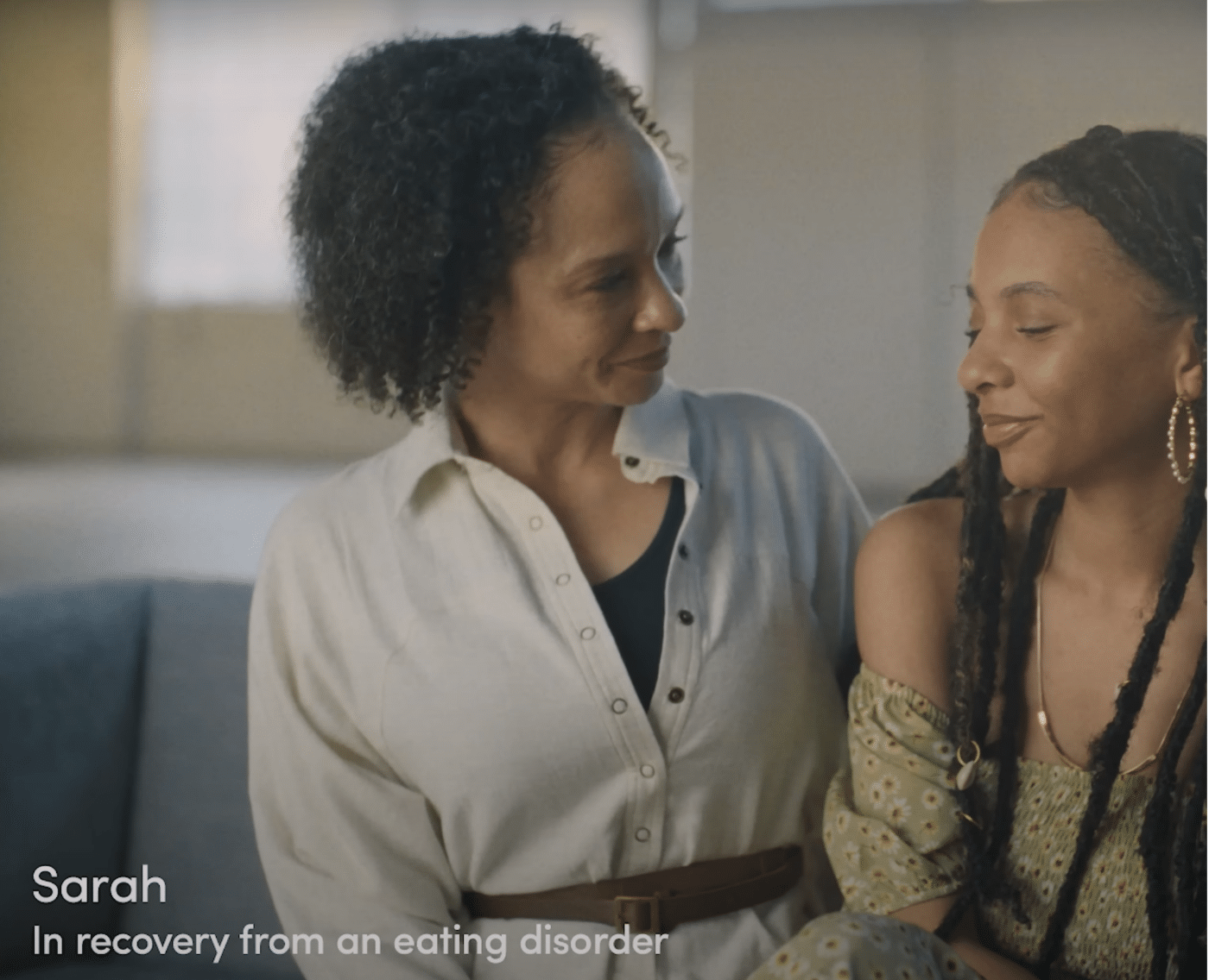 A frame from Dove’s campaign film, titled The Cost of Beauty. This image shows a tender moment between a mother and a daughter sitting close together. The woman on the left has curly dark hair and is wearing a light-coloured button-up shirt with a brown belt. She is looking at the other woman with a caring and supportive expression. The woman on the right has long braids, is wearing hoop earrings, and a green patterned off-shoulder top. She is smiling gently with her eyes closed, suggesting a sense of comfort or relief. The text overlay on the image reads, "Sarah - In recovery from an eating disorder".