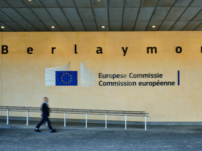 The Berlaymont building. A close-up of the European Commission lettering which sits to the right of European Commission logo, in the middle of the image. A man walks past in a blur underneath the logo.