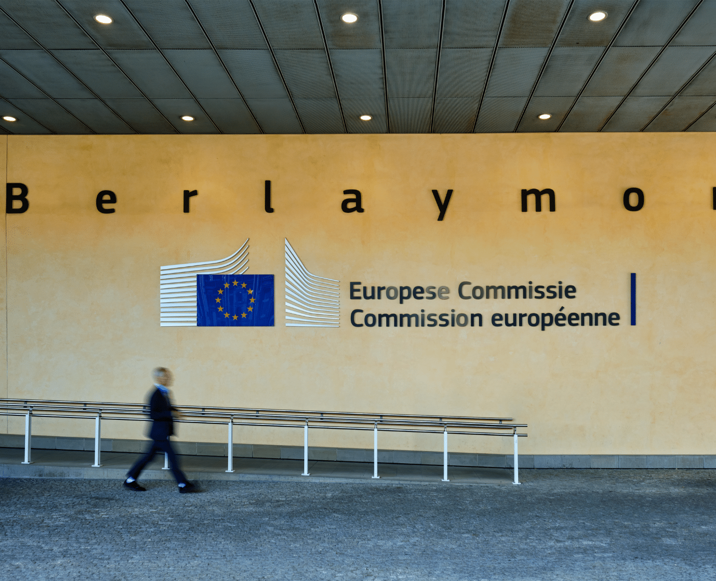 The Berlaymont building. A close-up of the European Commission lettering which sits to the right of European Commission logo, in the middle of the image. A man walks past in a blur underneath the logo. 