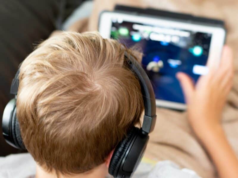 Boy in a white shirt playing a game on a white tablet computer while sat on a sofa.