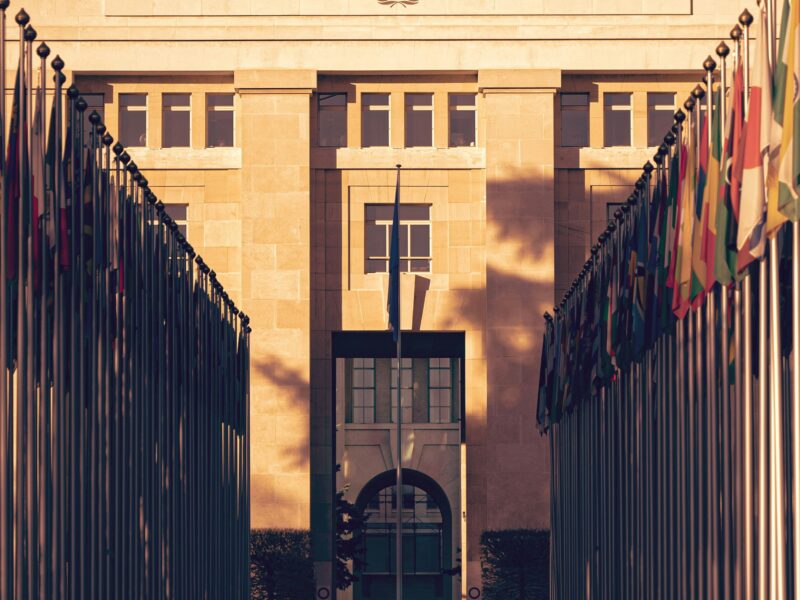 The Palais des Nations UN Headquarters in Geneva has a golden glow suggesting the photo was taken in early morning or late evening. The building fills the majoirty of the frame, with a strip of grey sky at the top of the frame. There is a dark low-level closed gate in the forground, and a row of flags leading to the building. All flags are drooped down, and caught in the shadow of trees.