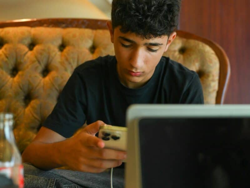 A teenage boy is sitting on a light green sofa. He is looking down at the smartphone in his right hand, and the smartphone is plugged into a charger. He face is showing limited expression, and the overall feel of the image is sad.