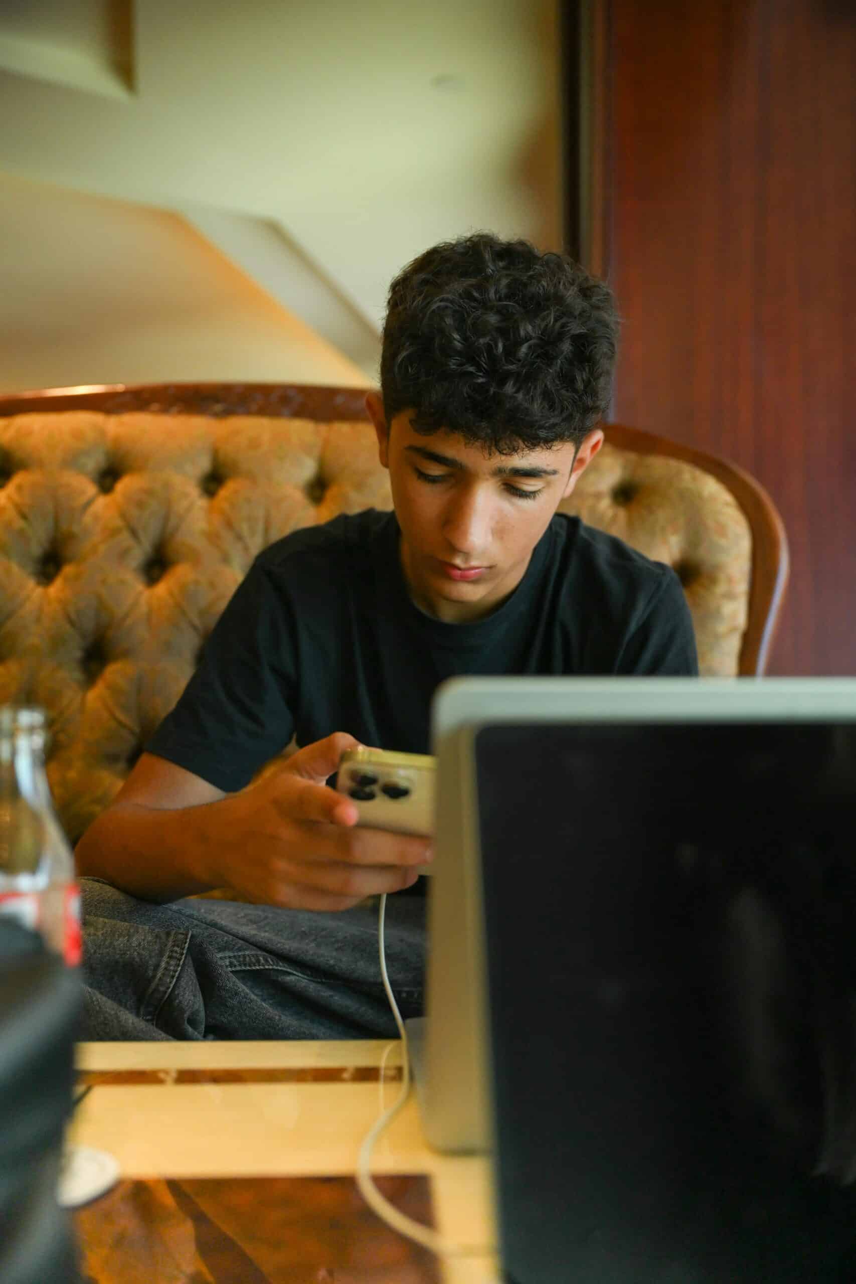 A teenage boy is sitting on a light green sofa. He is looking down at the smartphone in his right hand, and the smartphone is plugged into a charger. He face is showing limited expression, and the overall feel of the image is sad.